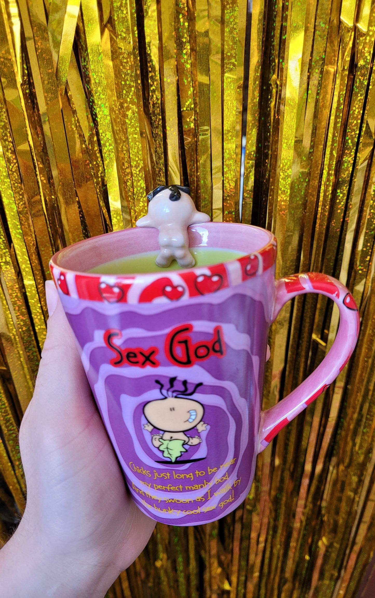 Mug with the 'sex god' figure hanging onto the rim of the mug. The mug has a red and pink handle with hearts on it and a groovy dark purple and lilac design on the main part of the mug
