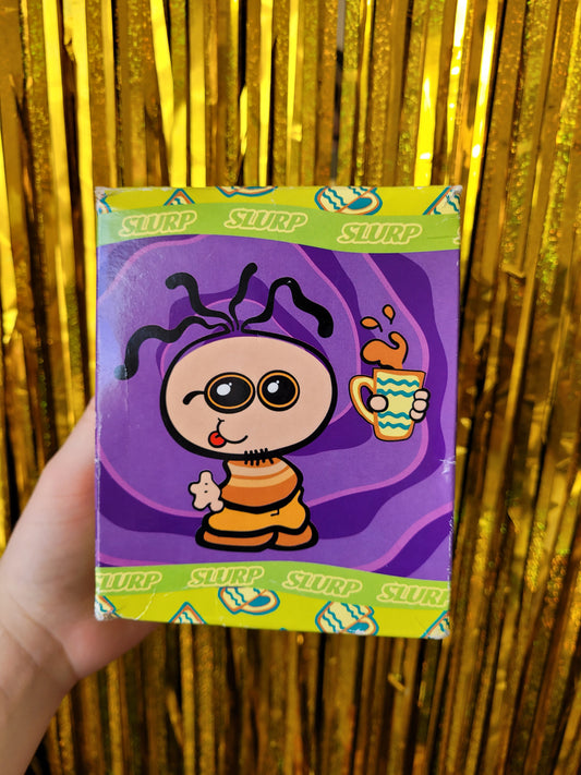 Side of the box of a cartoon man with a goatie beard and wiggly black hair. He's holding a cup of tea that is sloshing around and wearing and orange vest. The figure has sunglasses and is sticking his tongue out. The box has a groovy purple pattern and a green border that says 'SLURP' on repeat