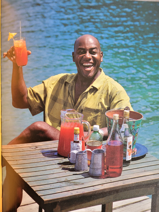 Ainsley sat at a table next to the sea holding a colourful drink with a cocktail umbrella and smiling at the camera