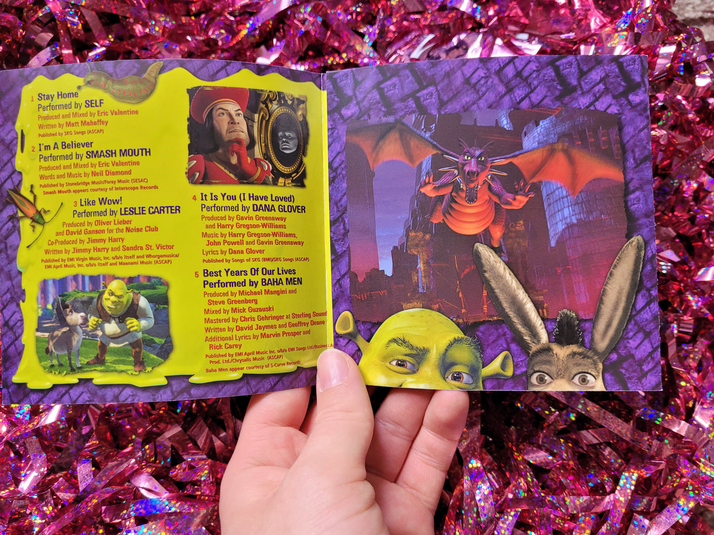 The opened booklet from inside the CD case with the track list surrounded by colourful pictures of scenes from the film such as Lord Farquaad plotting in front of the magic mirror and Dragon roaring and flying infront of a castle. 