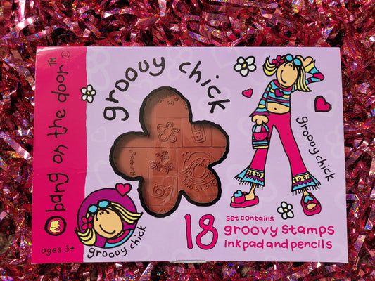 Closed box for the Groovy Chick stamps. The box is bright fuchsia pink and lilac with decorative daisies and pink hearts. It has the iconic 'groovy chick' figure on the front which is naively drawn cartoon of a girl with blonde hair flicked up at the bottom, sunglasses on her head, a colourful stripy top and flared pink jeans and sandals. The box reads 'Set contains 18 groovy stamps ink pad and pencils' The card box has a cellophane window where you can view the red rubber stamps. 