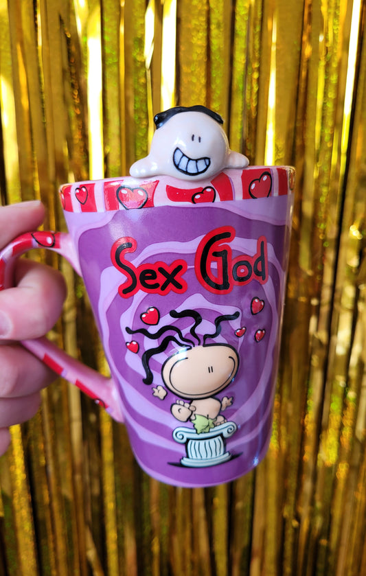 Purple and red mug saying 'Sex God' on the front in black and red curvy font above a round cartoon figure with wiggly black hair. The figure is naked apart from a strategically placed fig leaf. The character is standing on a roman style plinth surrounded by red love hearts. The rim of the mug has a 3D sex god figurine peeking over the side, as part of the mug.