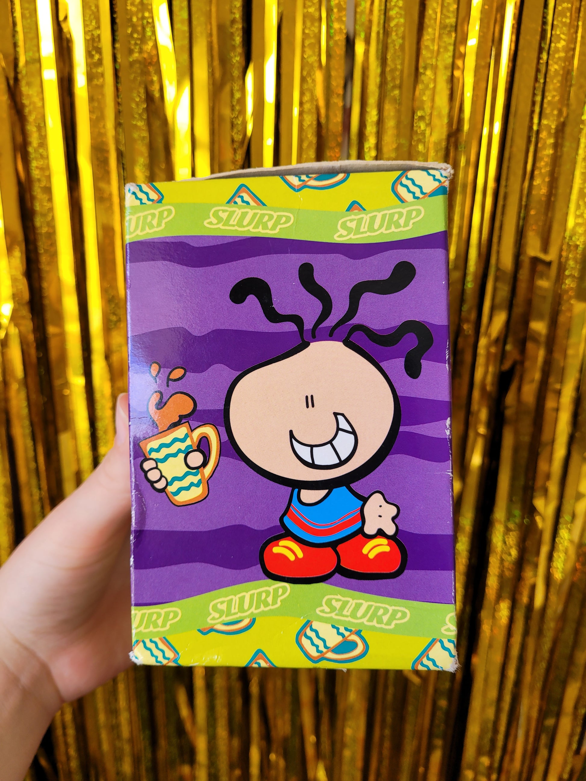 The other side of the box with a different character holding a mug of tea sloshing about. The character has a blue and red sports vest and red and yellow oversized trainers and is grinning.