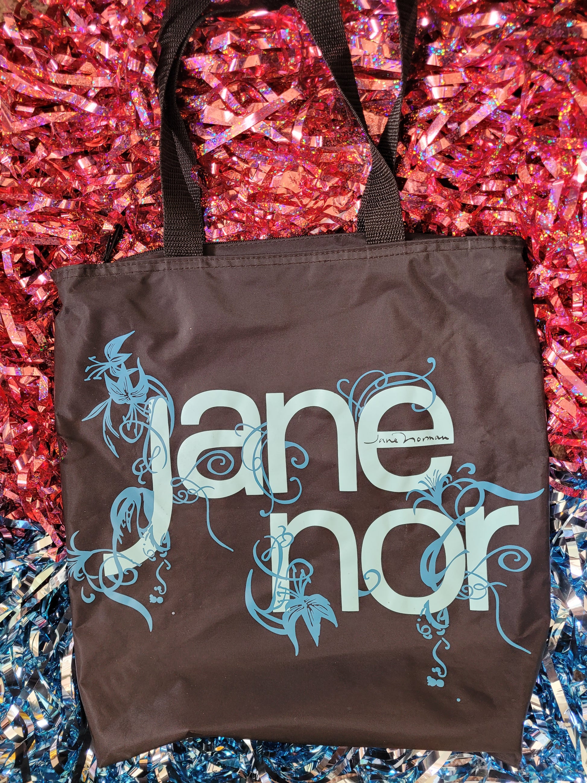Black tote style bag with black handles. The bag has large rounded font saying 'Jane Norman' covering most of the front of the bag. The light blue lettering is surrounded by a darker blue flowers and leaves swirling decoratively around the letters. Witihin the 'e' of the 'Jane' reads the black Jane Norman logo 
The bag is against a two tone pink and blue sparkly tinsel background