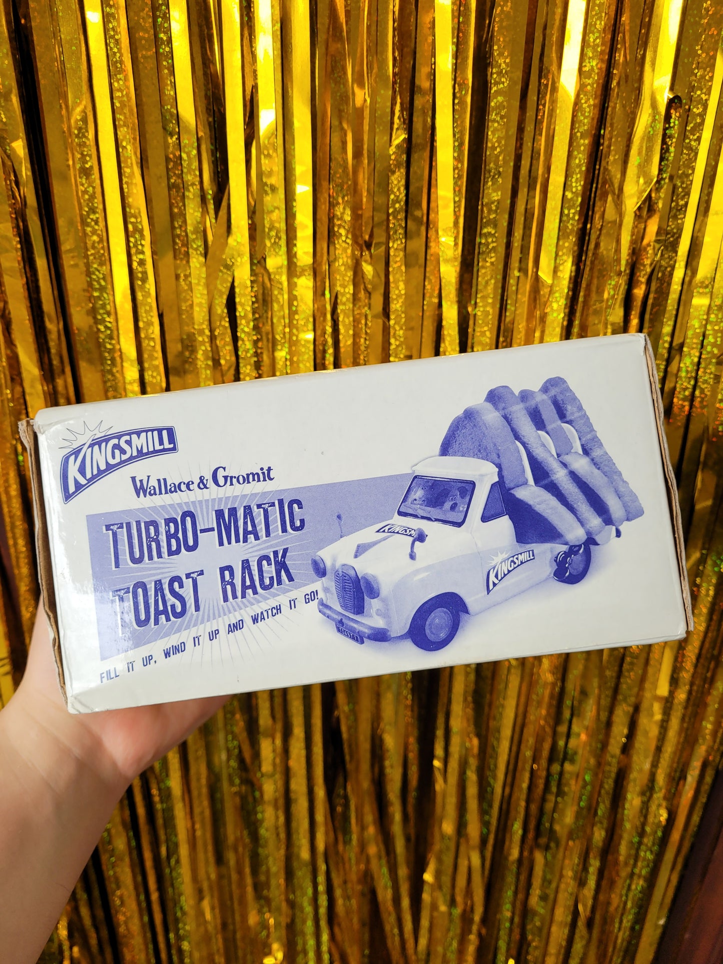 Rectangular cardboard box reading 'Kingsmill Wallace & Gromit Turbo-Matic Toast Rack' in dark blue all caps letters against a white background 'Fill it up, wind it up and watch it go!' with a picture of the toast rack with slices of toast in