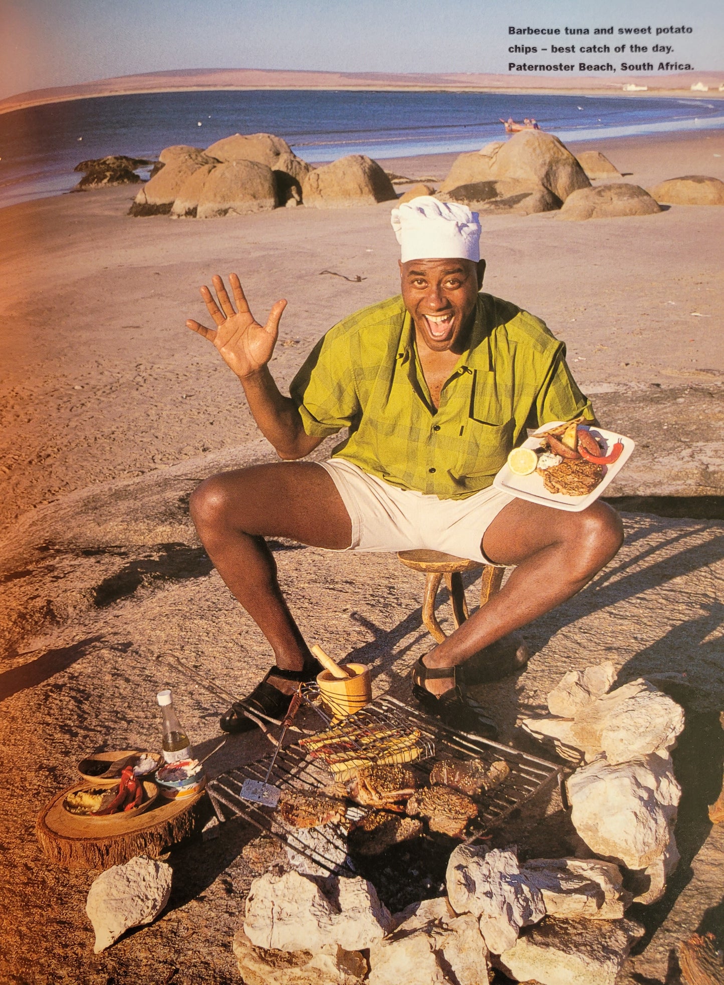 Ainsley wearing a chefs hat, sat on a sandy beach in front of a barbeque, holding a plate of food and smiling towards the camera.