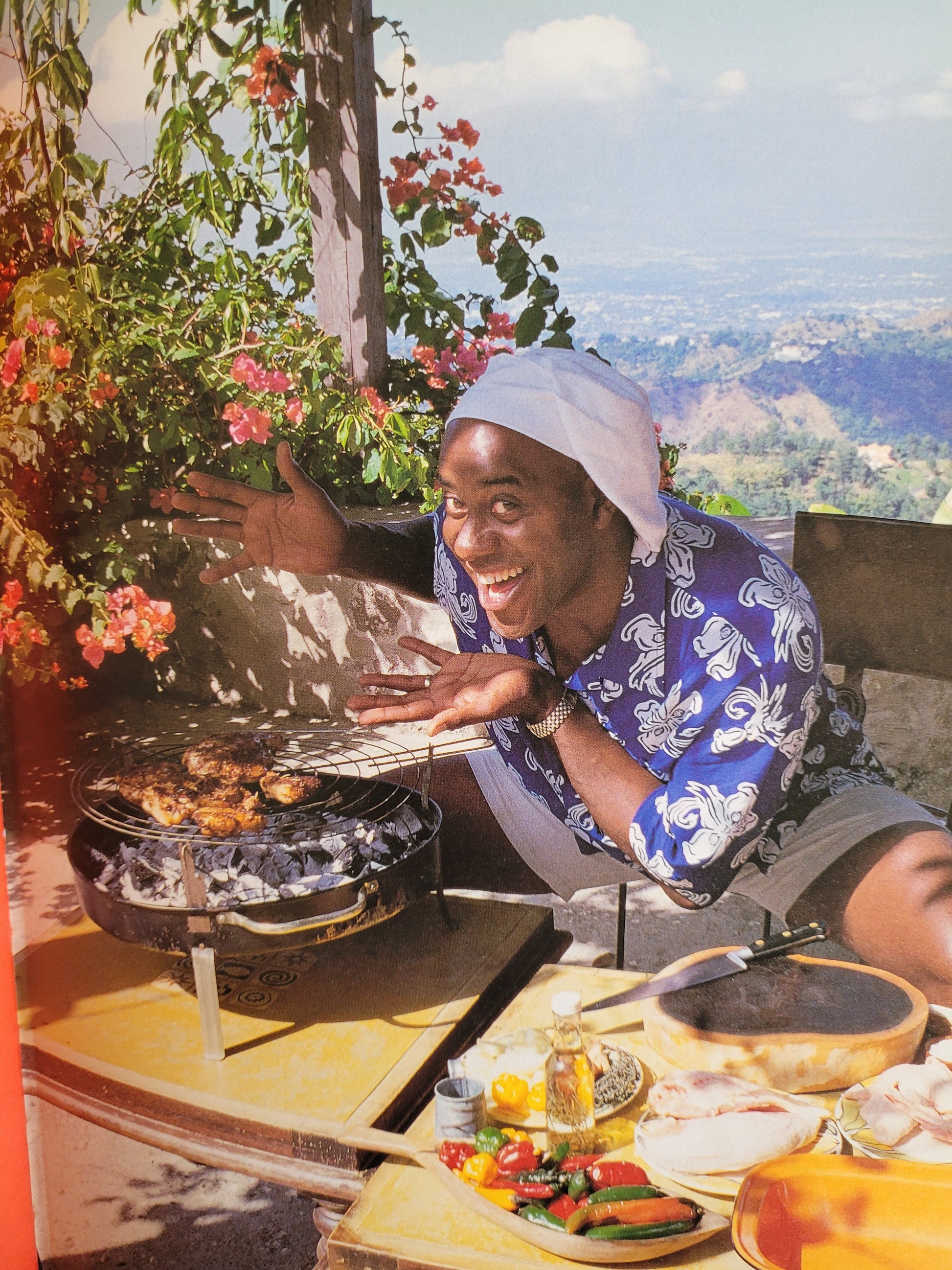 Ainsley wearing a blue and white hawaiian shirt smiling enthusiastically towards the camera, gesturing towards some chicken grilling on a barbeque. Ainsley is sat in front of a plant with bright pink flowers and the backdrop is a view of grass, trees and hills. 