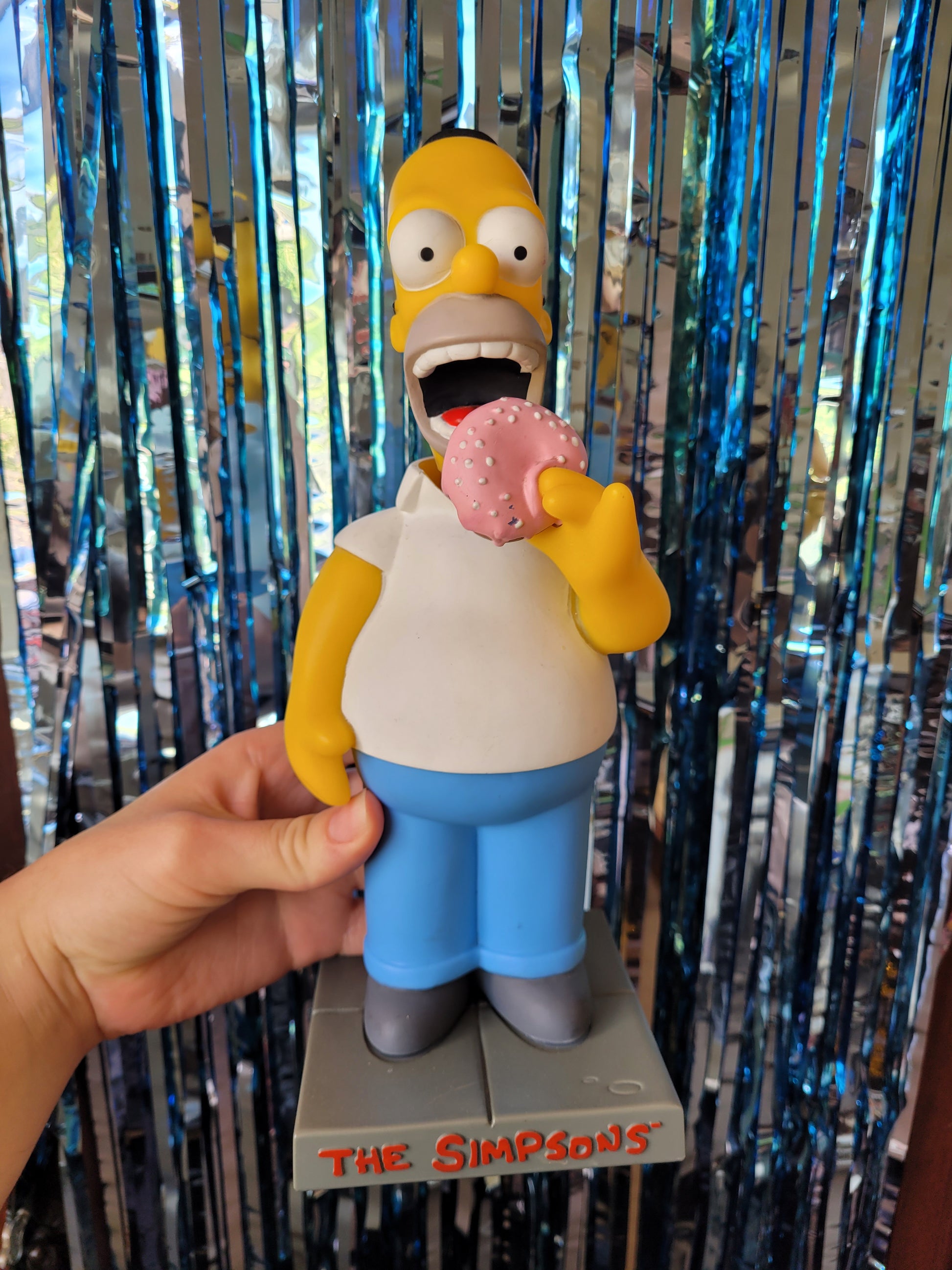 Plastic figure of Homer Simpson bobblehead eating a pink and white donut on a small grey platform with The Simpsons TM in red writing across the front. Picture taken against a sparkly blue tinsel background