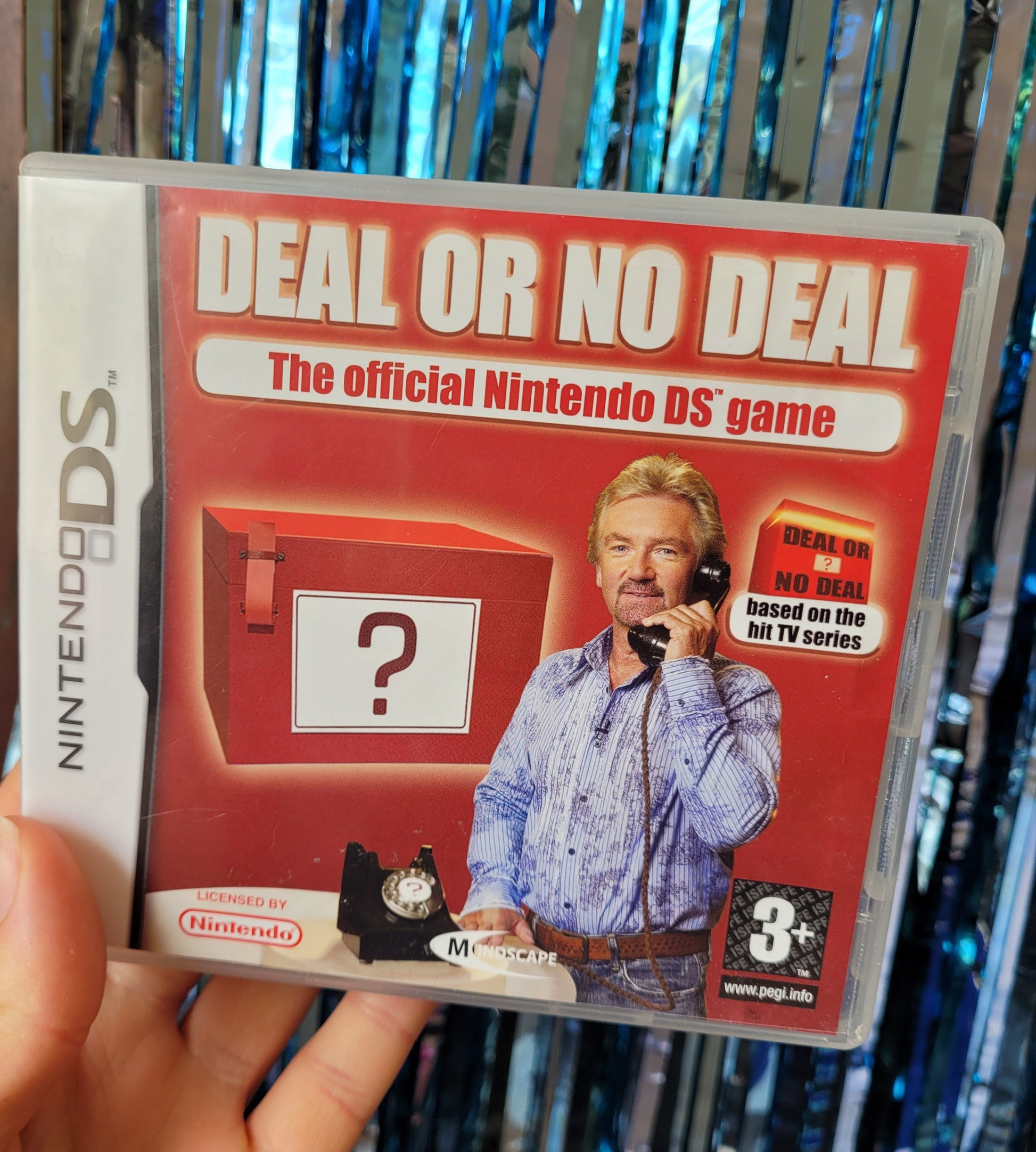 Deal or No Deal Nintendo DS Game case with a picture of Noel Edmunds wearing a striped and floral blue shirt. He is smiling at the camera and holding the banker's phone to his ear standing in front of a picture of a Deal or No Deal box with a '?' on it 