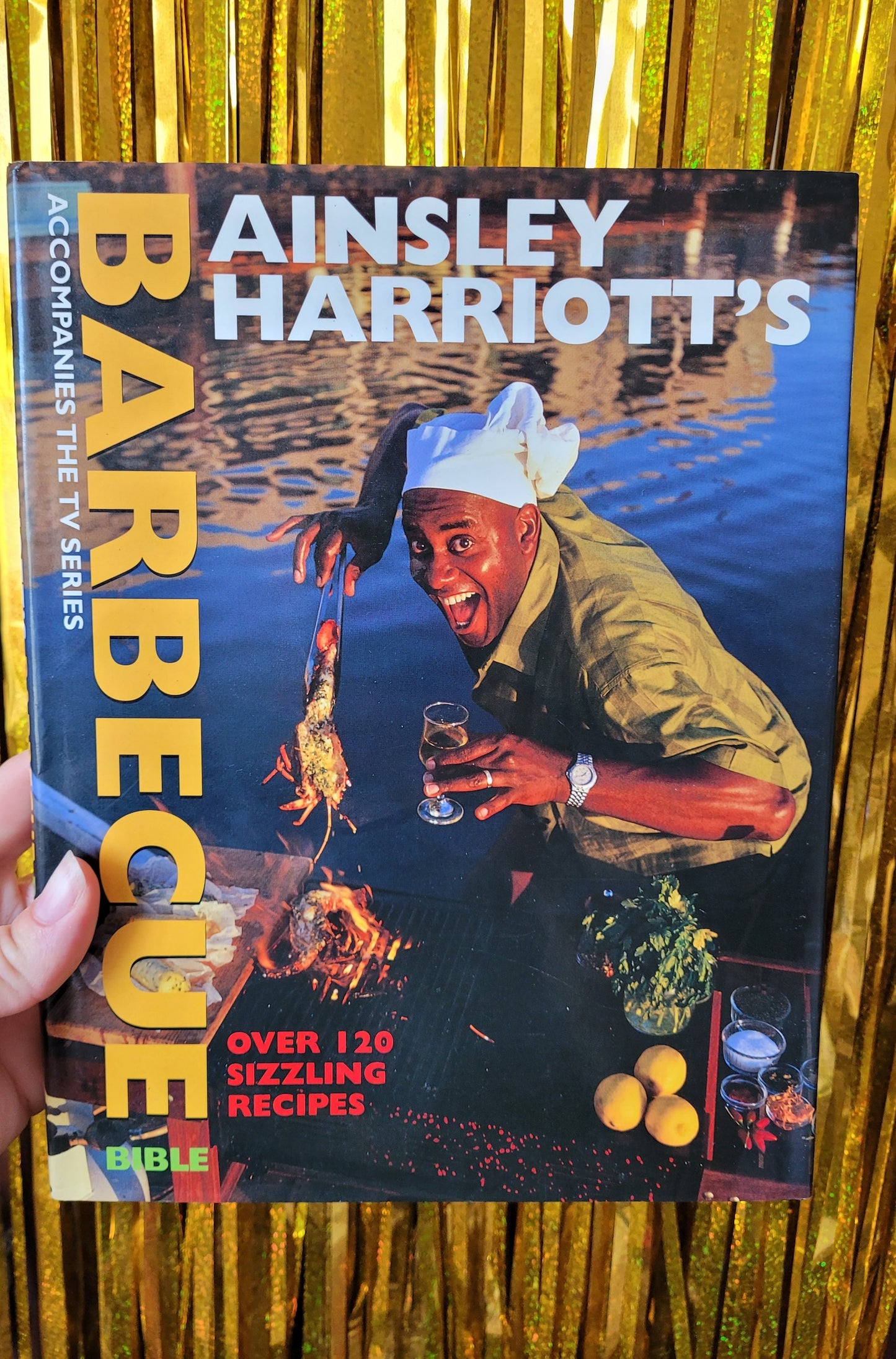 Frontcover of book titled 'Ainsley Harriorr's Barbeque Bible' 
'Over 120 Sizzling Recipes' 
'Accompanies the TV Series'

The colourful picture on the front cover is Ainsley smiling enthusiastically at the camera, wearing a chef's hat holding up some barbequed seafood and holding a glass of wine stood in front of the sea

The photo of the book is taken in front of a sparkly gold tinsel backdrop 