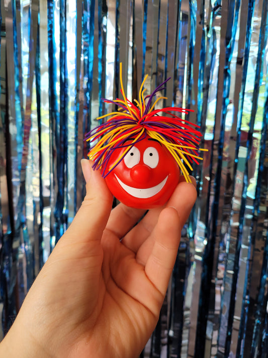 Plastic fancy dress red Nose accessory with grinning expression and a bright yellow, purple and red elastic hair do.  Item against blue sparkly tinsel background.