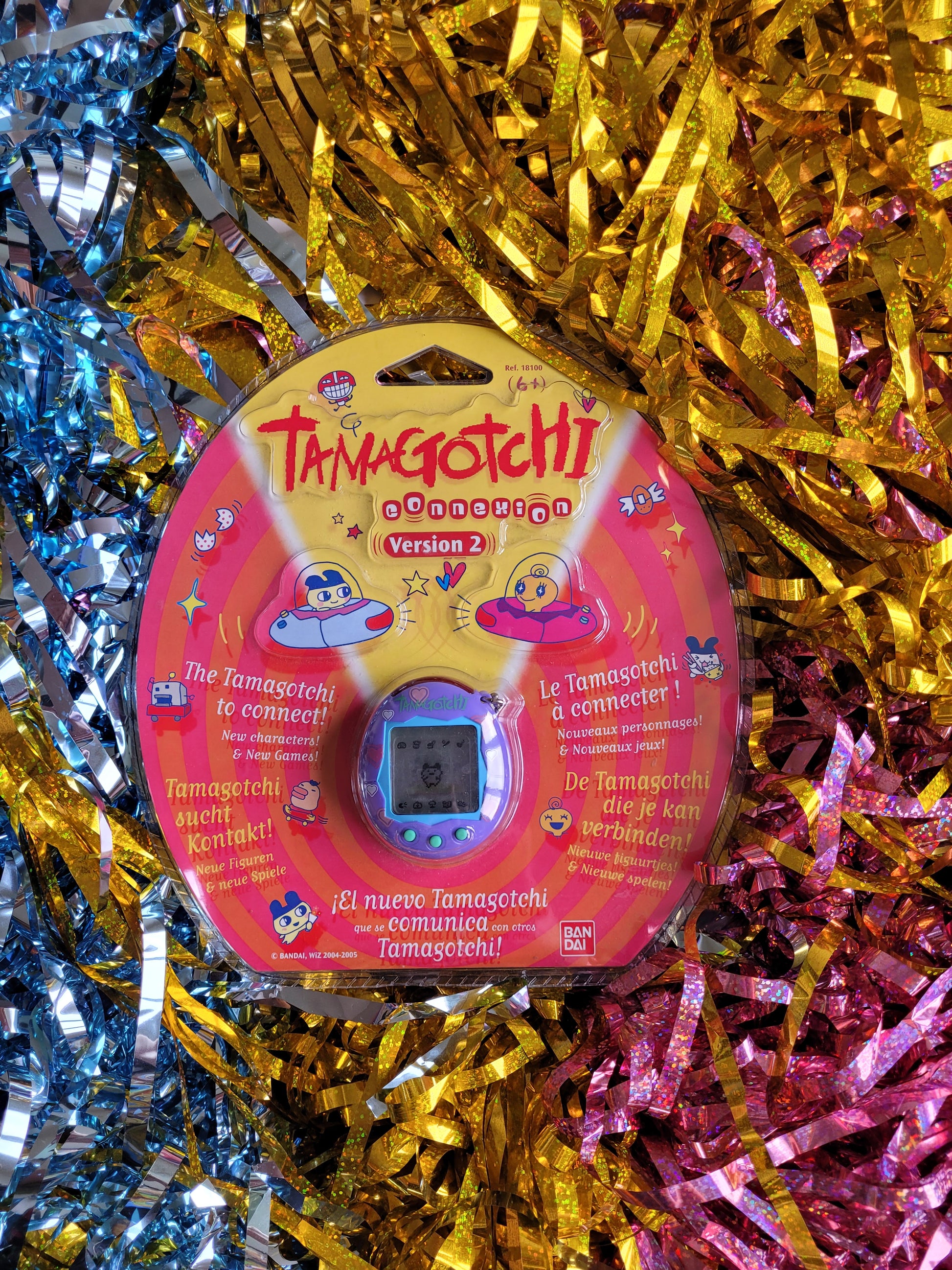 Purple and pink tamagotchi in plastic packaging against multicoloured tinsel background