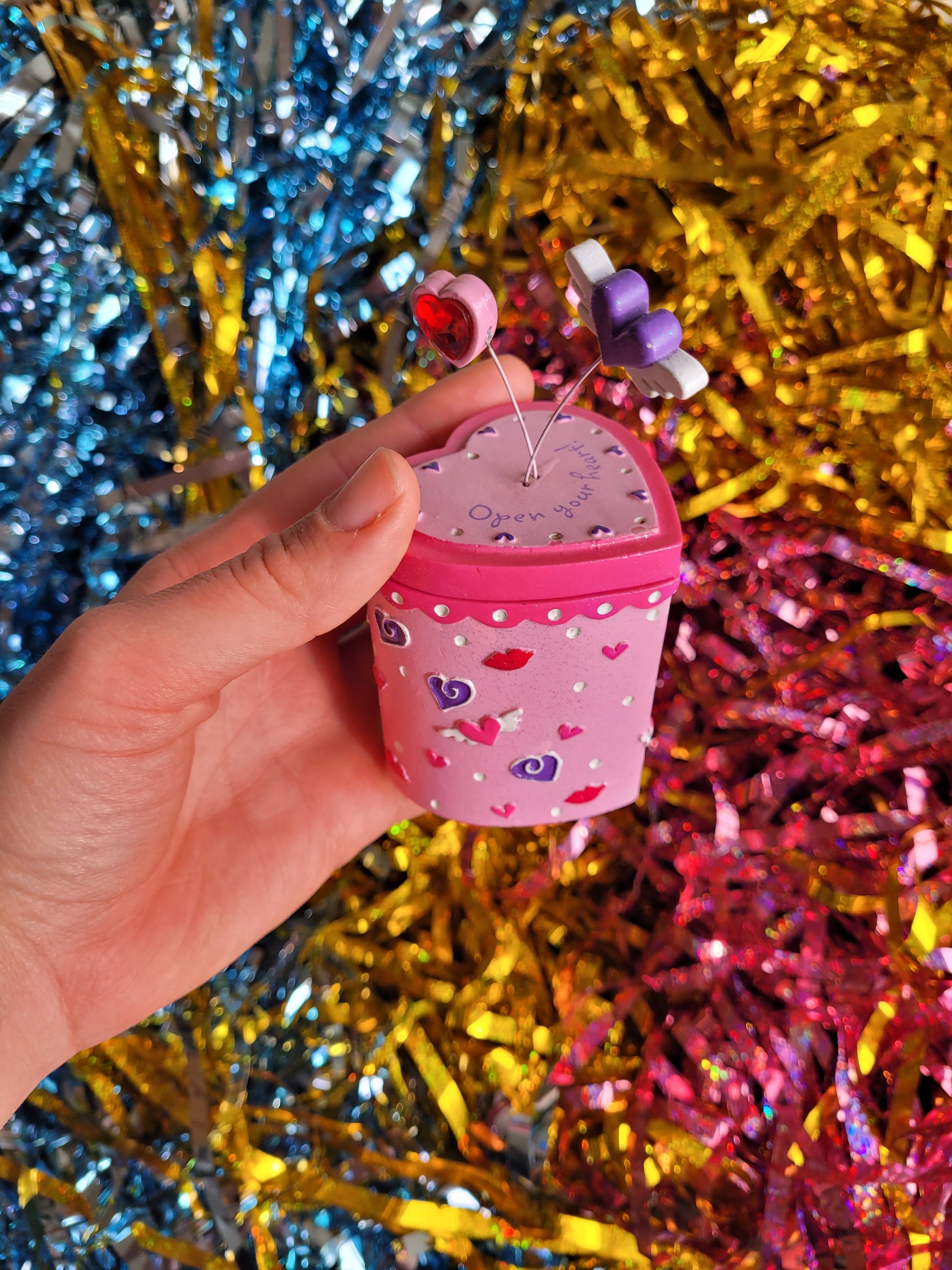 Pink and purple trinket box with heart pattern. Springing out the top of the lid are two hearts attached by wire. The top of the lid reads 'open your heart' 