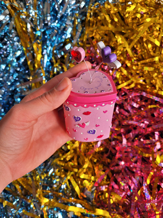 Pink and purple trinket box with heart pattern. Springing out the top of the lid are two hearts attached by wire. The top of the lid reads 'open your heart' 