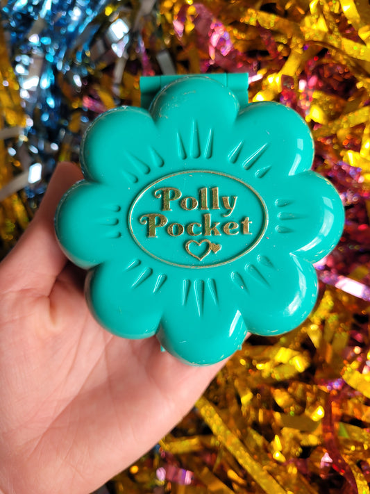 Turquoise plastic polly pocket case in the shape of a flower against a multicoloured tinsel background 