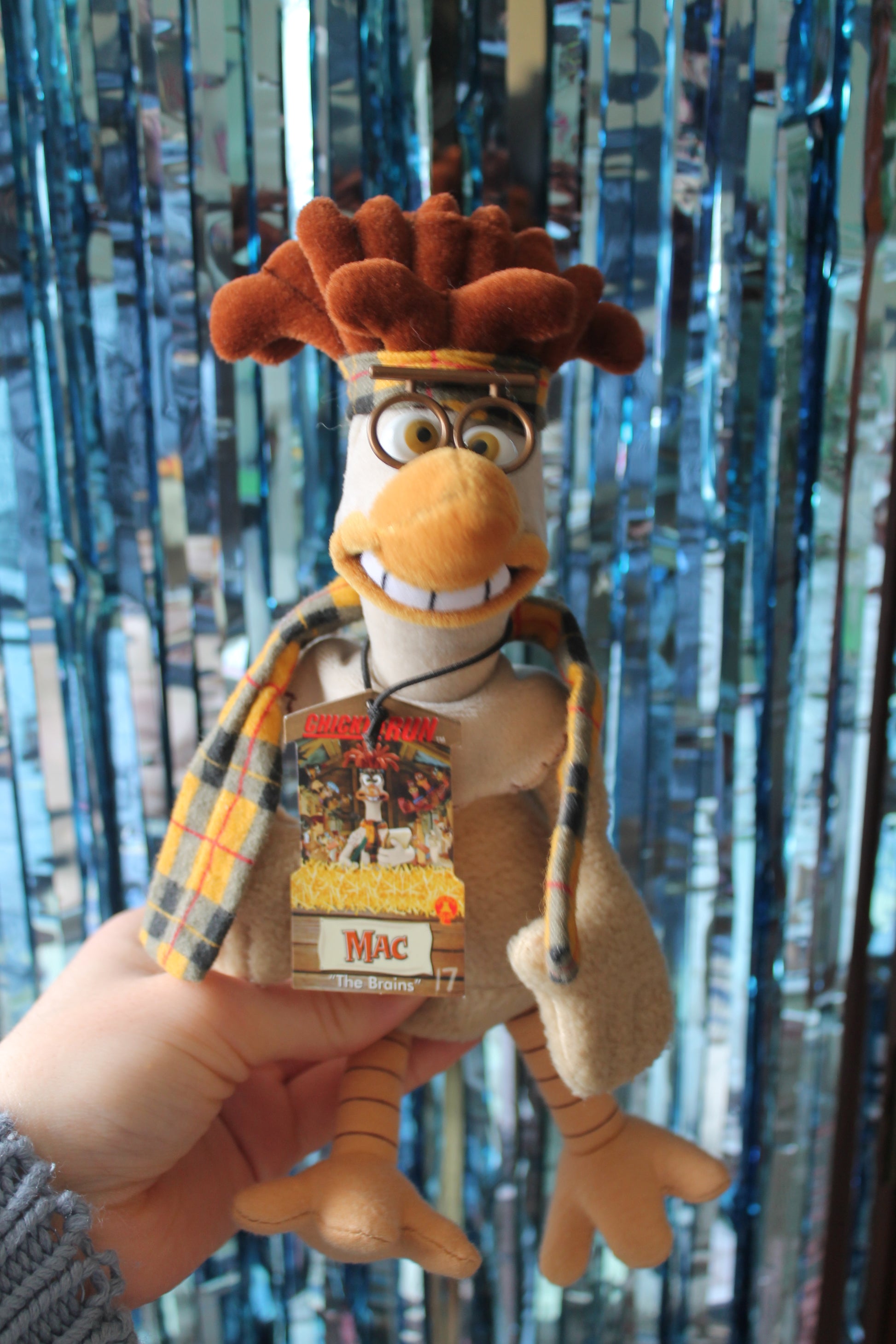 Plush beige, brown and yellow toy. The character is a chicken with round glasses, teeth and a tartan headband and scarf. The toy is handheld in the photo against the backdrop of a sparkly blue foil curtain.