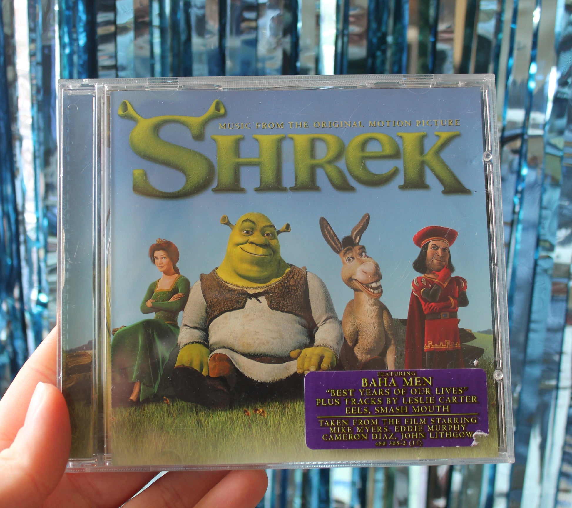 Shrek CD case held in front of a blue sparkly tinsel background. The CD case has 'Shrek' in large green letters at the top. The 'S' has Shrek's elongated tubular ogre ears attached. The image on the CD cover is Princess Fiona with her arms folded smiling and looking slightly fed up. Shrek with his eyebrow raised knowingly. Donkey with a open mouthed grin looking jovial and Lord Farquaad stroking his chin looking dastardly. All stood or sat on grass with a clear blue sky background