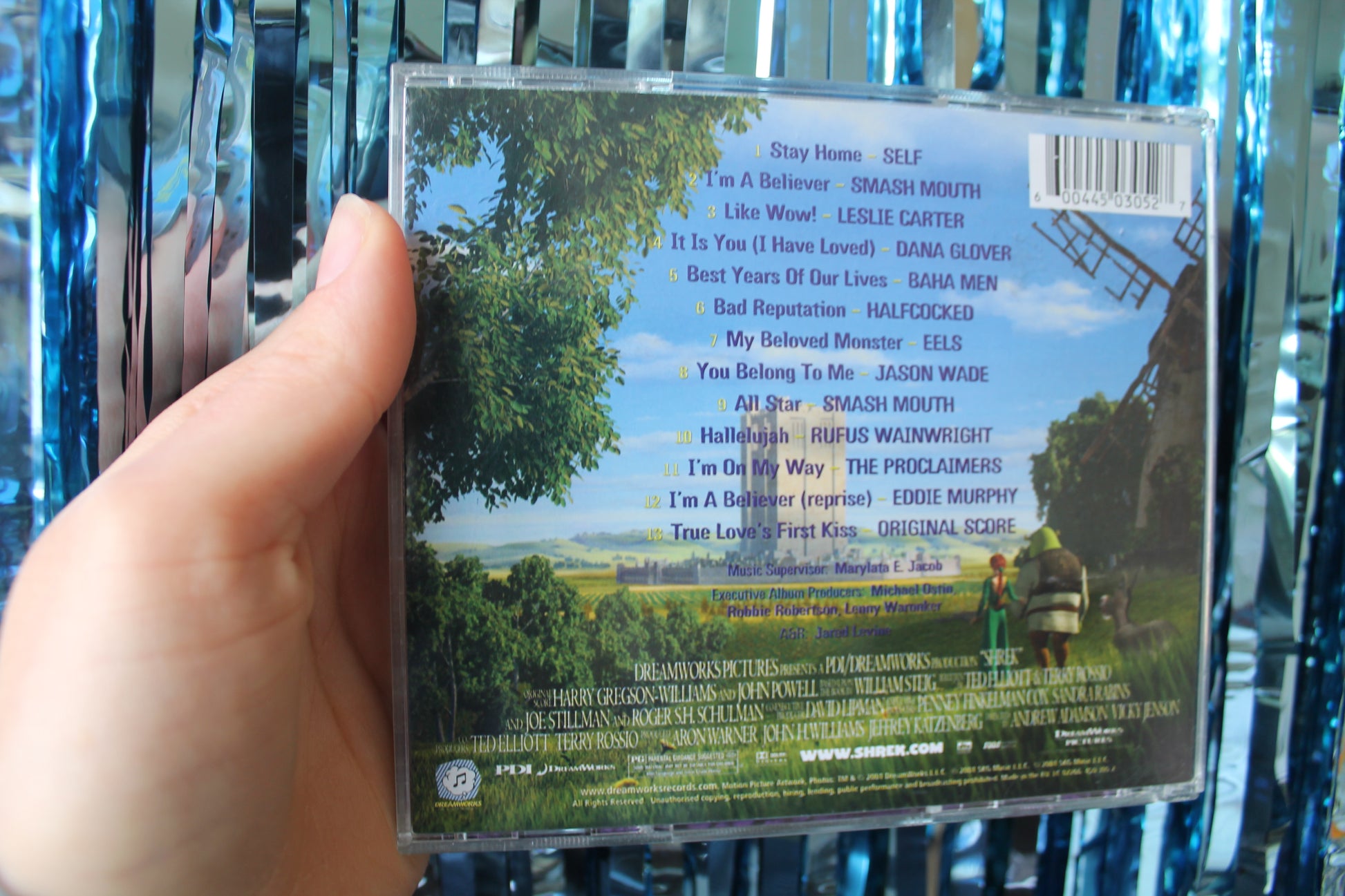 The back of the CD with the track list against a scenic background of trees and grass with a large grey rectangular castle in the middle. 