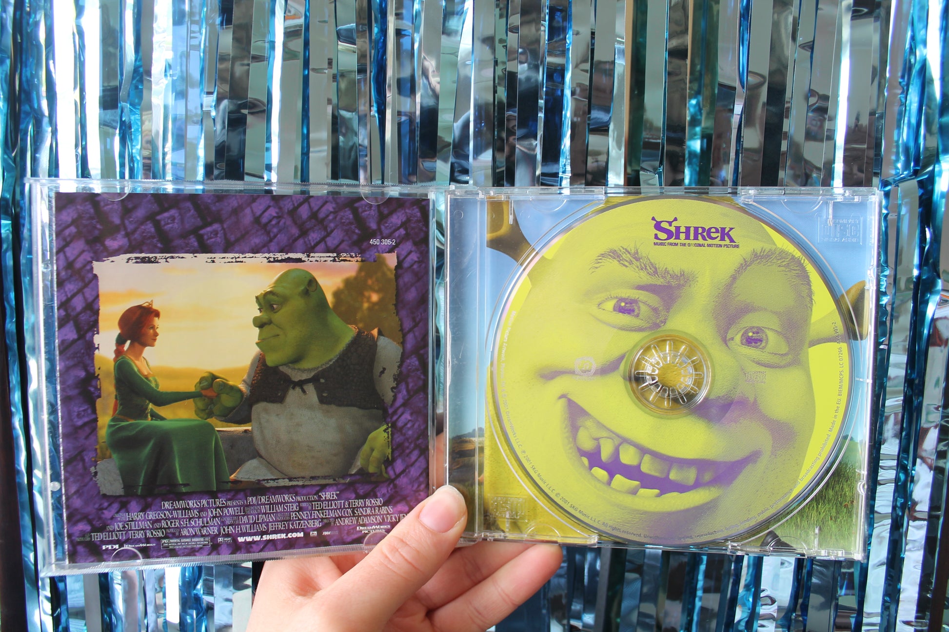Shrek CD case held open in front of a blue sparkly tinsel background. The back of the CD booklet has a picture of Shrek and Princess Fiona looking at each other adoringly and holding hands.