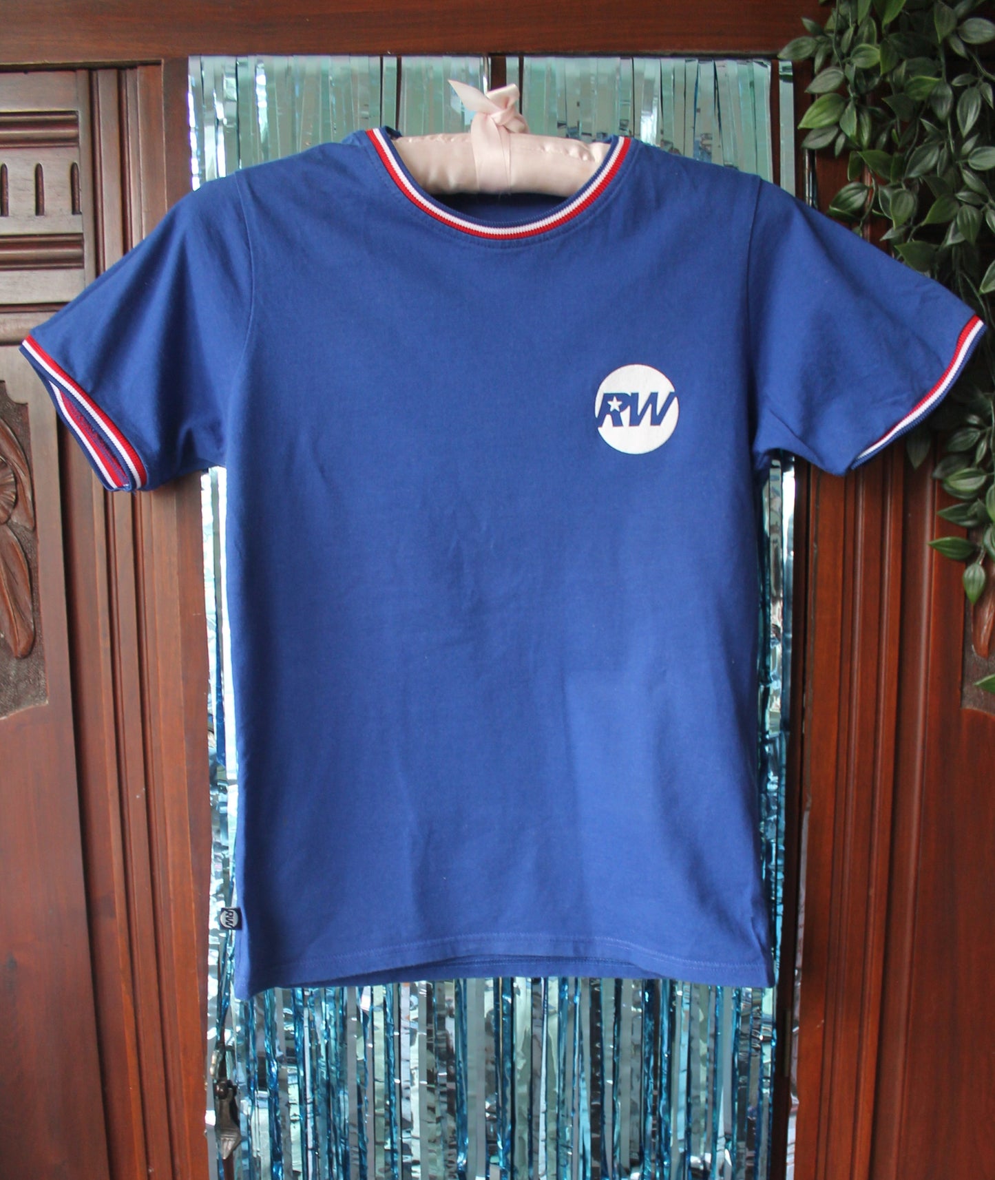 Blue football shirt with red and white hem on the sleeves and collar. Official white RW logo on the front of the shirt. Against a blue, sparkly tinsel background. 