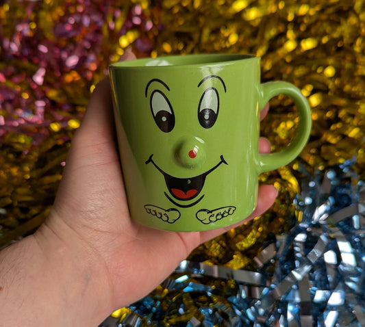 Small green mug with a smiling face and a button shaped nose that sticks out 